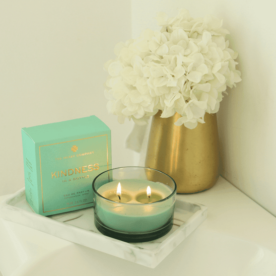 Kindness in a Bottle by The Heart Company displayed on a marble tray beside a lit candle and white flowers in a gold vase, creating a calm, elegant atmosphere that reflects a vegan, cruelty-free unisex fragrance with a fresh, woody scent for self-care and mindful moments