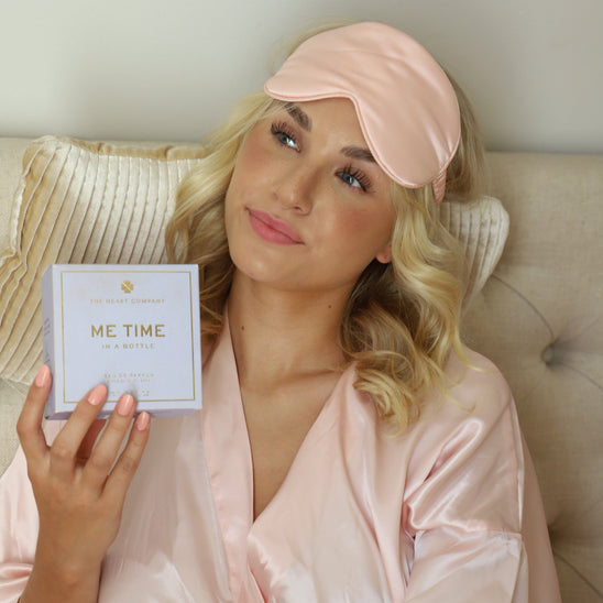Woman in pink robe and sleep mask holding Me Time in a Bottle vegan lavender perfume box by The Heart Company, relaxing self-care moment with elegant fragrance packaging
