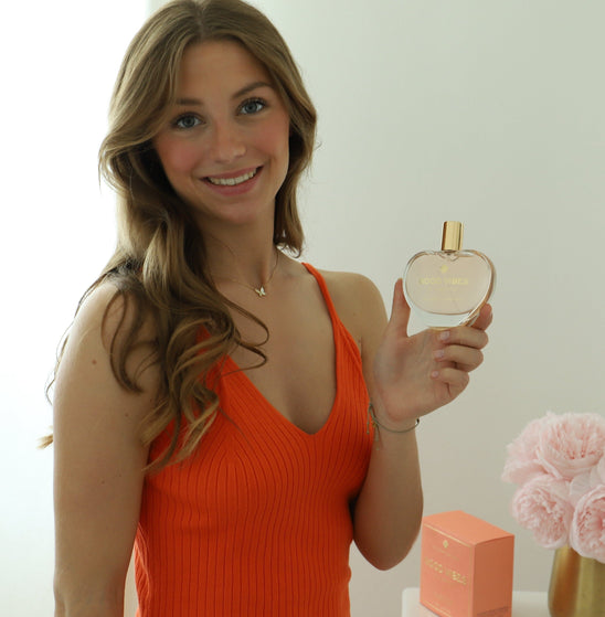 Smiling woman holding “Good Vibes in a Bottle” vegan eau de parfum by The Heart Company, heart-shaped perfume bottle with gold cap, fruity floral sweet gourmand fragrance, cruelty-free luxury perfume for positive energy and everyday wear