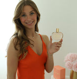 Smiling woman holding “Good Vibes in a Bottle” vegan eau de parfum by The Heart Company, heart-shaped perfume bottle with gold cap, fruity floral sweet gourmand fragrance, cruelty-free luxury perfume for positive energy and everyday wear