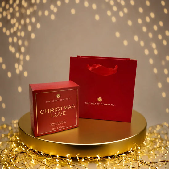 Christmas Love vegan eau de parfum by The Heart Company displayed with red gift box and branded shopping bag on a gold platform with festive lights, cruelty-free holiday fragrance gift