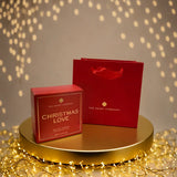Christmas Love vegan eau de parfum by The Heart Company displayed with red gift box and branded shopping bag on a gold platform with festive lights, cruelty-free holiday fragrance gift