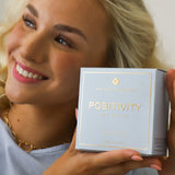 Smiling woman holding Positivity in a Bottle by The Heart Company, a vegan eau de parfum in a light blue box with gold accents, representing a fresh, aquatic, clean fragrance that is paraben-free and phthalate-free, designed to uplift mood, inspire positivity, and support everyday wellness